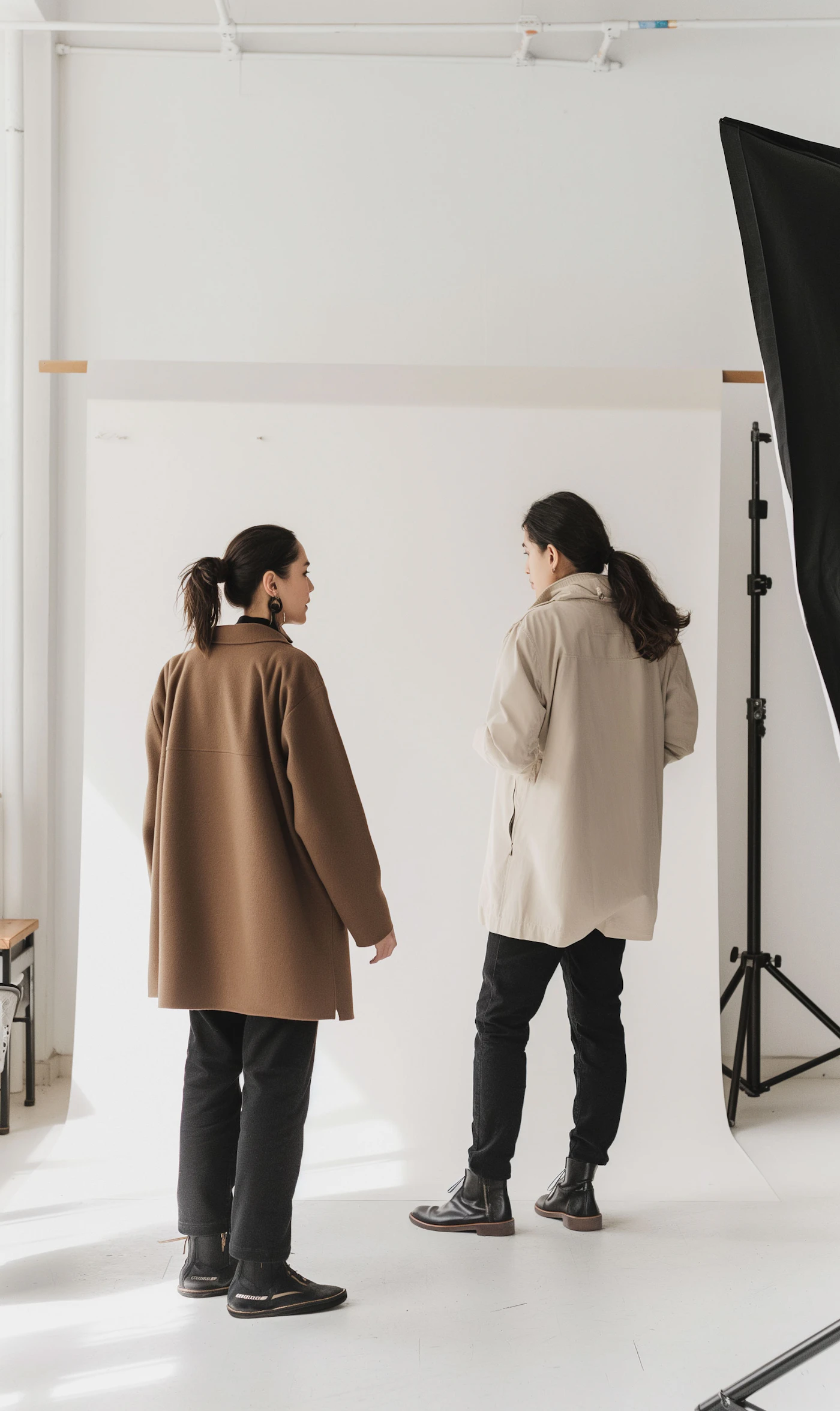 Two people in long coats talking on a bright studio set beside the lighting rig